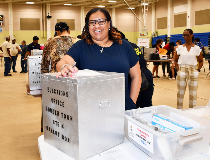 Image of a smiling woman holding a paper ballot over a ballot box inside a polling station in the Cayman Islands, during a previous Election Observation mission delivered by CPA BIMR.